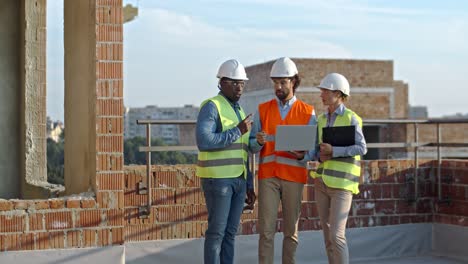 Caucasian-woman-and-mixed-races-men-standing-together-in-hardhats-at-the-building-site-with-laptop-computer,-workday-of-builders,-constructors-and-architects.
